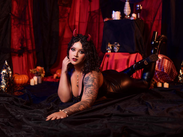 ValeryGotti with curly black hair, devil horns, black leather outfit, tattoos, lying on black fabric, hand on cheek, boots, red and black background, candles, pumpkins, and a table.