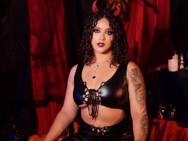 ValeryGotti with curly black hair, red devil horns, black leather crop top with lace-up front, showing her tits, tattooed arm, and red lipstick, in a dark, red-lit setting.