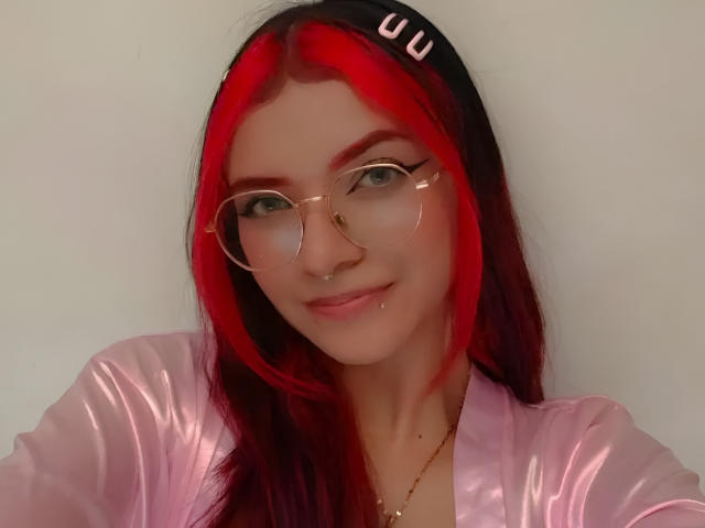 AriaRosel with red and black hair, wearing glasses, a pink shiny top, nose ring, and lip piercing, smiling softly, has a small gold necklace.