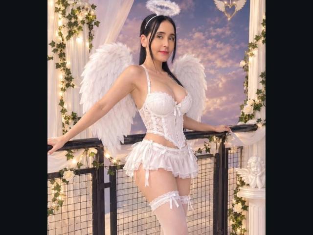 MindyLove con ali bianche da angelo, un'aureola e biancheria intima bianca, che mostra il suo seno e i suoi glutei, in piedi accanto a una ringhiera nera, indossando calze alte fino alla coscia e un corsetto in pizzo..