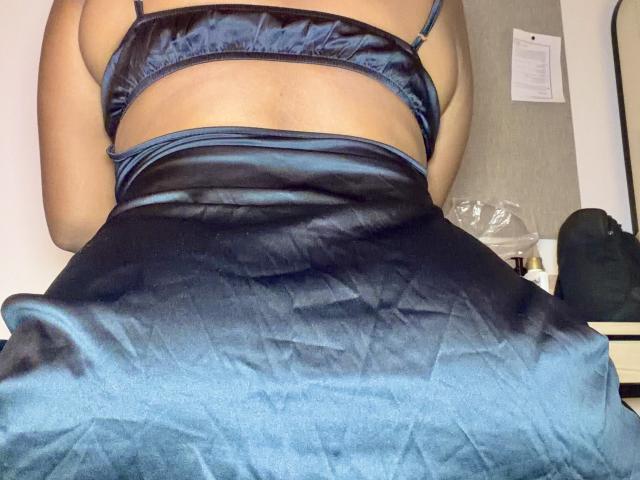 IvannaDuran69's back is to the camera, wearing a shiny black satin bra and matching skirt, showing her tan skin and curvy butt. Her skirt is slightly raised.
