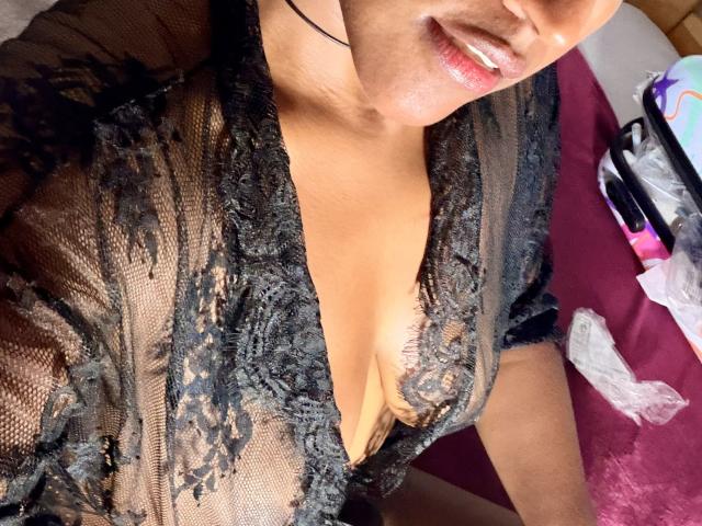 IvannaDuran69 wearing a black lace robe, revealing her cleavage and small tits, with a black choker and glossy pink lips, lying on a maroon bed.