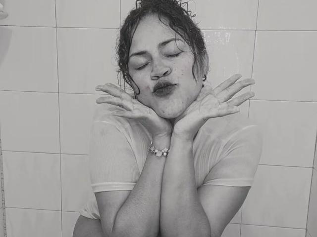 IvannaDuran69, wet shirt clinging to her body, hands up, pouty face, water dripping from her hair, eyes closed, freckles visible, wearing a bracelet, black-and-white photo.