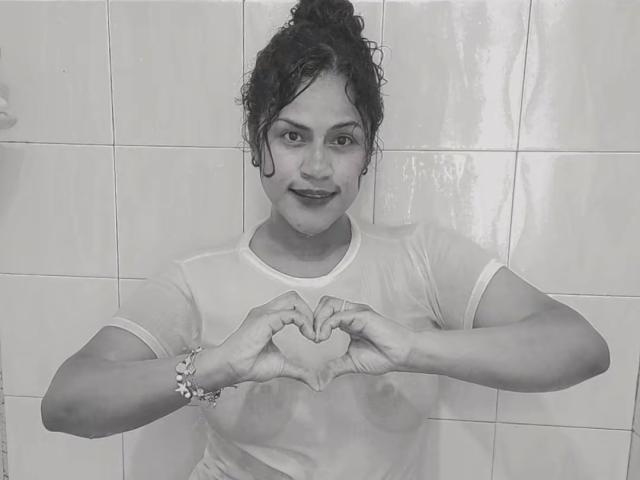 IvannaDuran69 making a heart shape with wet hands, wearing a see-through shirt, showing small tits, with curly hair, and a bracelet, in a black-and-white photo.