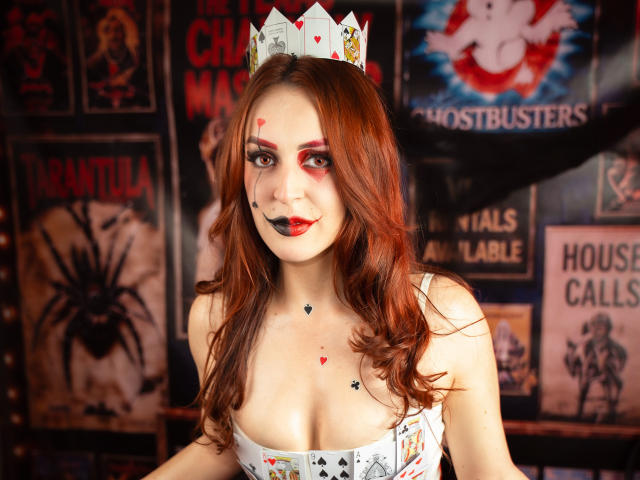 ButtyCherry with red hair, wearing a card crown, face painted with red lines and black heart marks, white dress with playing cards, showing tits, in a dark, spooky room.