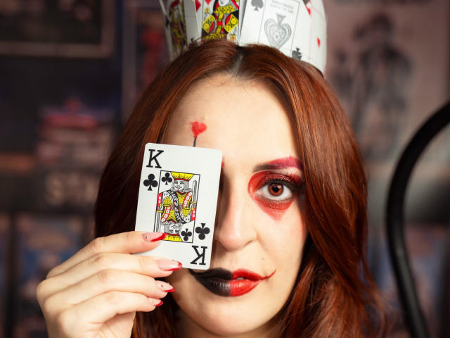 ButtyCherry with red hair, wearing a playing card hat, holding a King of Spades card over one eye, red makeup, lipstick, and nail polish, with a small red heart on cheek.