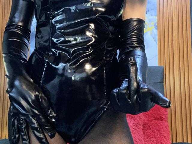 LilithStorm wearing shiny black latex dress and gloves, giving a thumbs-up with her right hand, showing off her ass and black panties, in a wooden-paneled room.