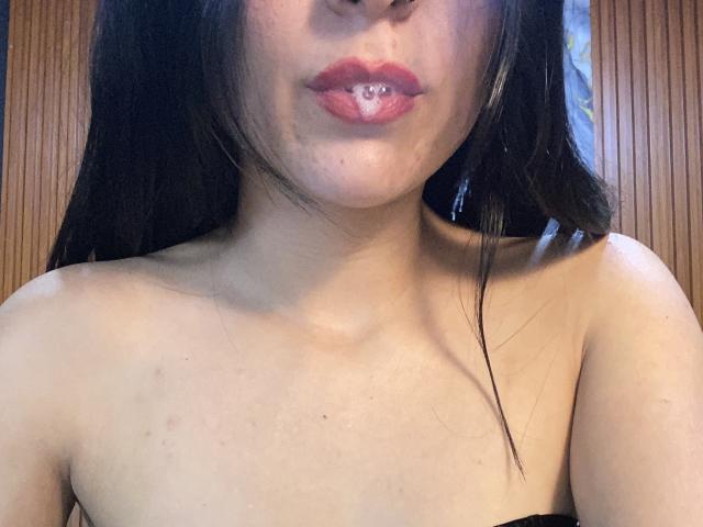 LilithStorm with dark hair, pink lipstick, and a small white liquid drop on her lower lip, bare shoulders, light skin, and a few small blemishes on her chest.