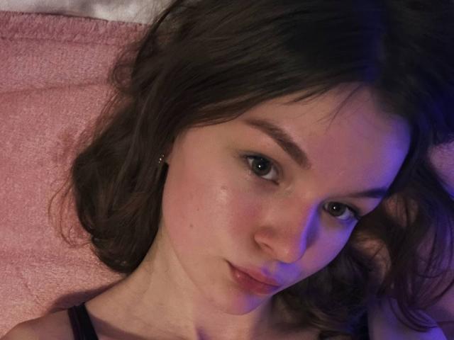 AlyssaVilliard with wavy brown hair, light skin, and subtle blue lighting, lying on a pink pillow, looking at the camera with a slight smile, showing her bare shoulders.