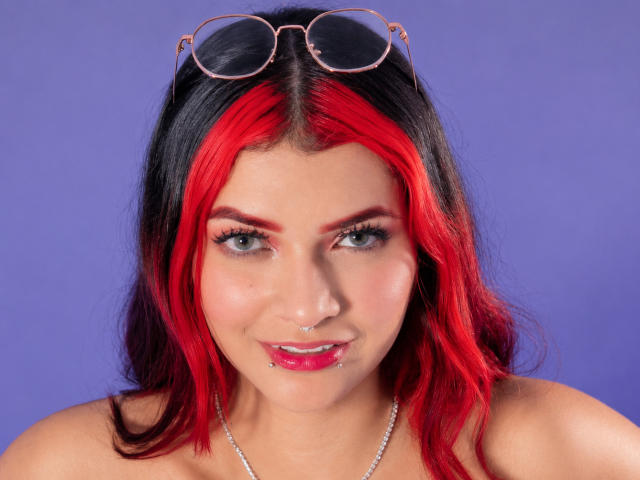 AriaRosel with red and black hair, blue eyes, pink lipstick, nose ring, and glasses on head, smiling at camera, wearing a silver necklace.