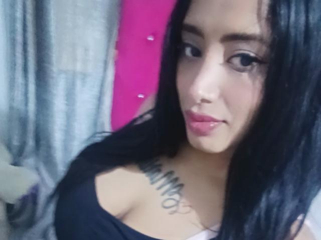 MariaCatalina with black hair, pink lips, and large tits, wearing a black top, showing a tattoo across her chest. She's looking at the camera with a slight smile.