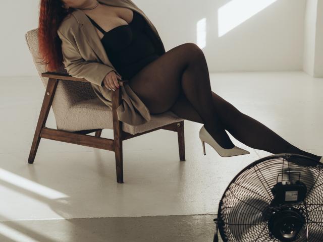 Dinastiia with red hair, wearing a beige jacket, black lingerie, and sheer pantyhose, sitting in a chair with legs crossed, white heels, and a standing fan. Boobs.