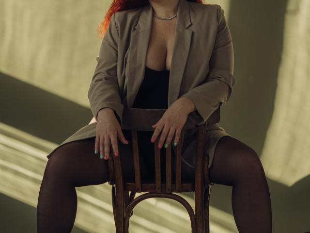 Dinastiia, avec ses cheveux roux, son blazer beige, sa robe noire et ses collants noirs transparents, assise sur une chaise en bois, les jambes écartées, montrant ses seins, avec des ongles peints en rose et en vert..