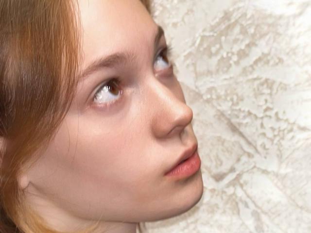 Il viso di Loni Tockey, con la pelle pallida, gli occhi marrone chiaro e le labbra rosa, con i capelli rossi, guarda di lato, sullo sfondo di una parete bianca con una texture..