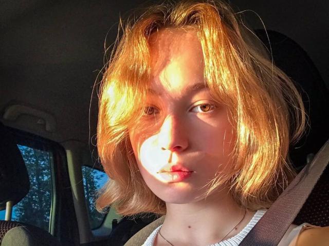 Loni Tockey, con i capelli biondi e ondulati, il viso illuminato dal sole, un'espressione seria, indossa la cintura di sicurezza, una camicia bianca e una collana, all'interno di un'auto. Il suo seno non è visibile..