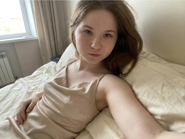 PamGillilan with freckles, wearing a beige tank top, lying on a bed, holding a phone for a selfie, showing a nose ring, with a relaxed expression.
