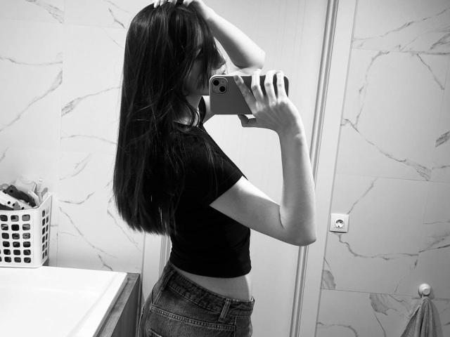 JinAvans with long dark hair, wearing a black crop top and jeans, takes a mirror selfie with her right hand in her hair, showing her side and midriff.