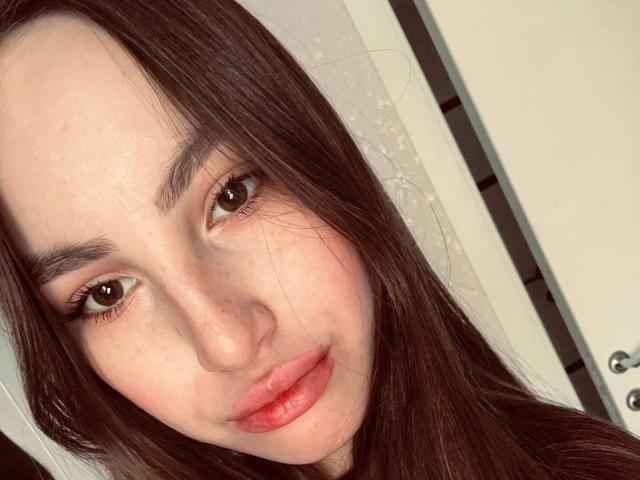 JinAvans has long brown hair, brown eyes, and pinkish-red lips with a slight pout. Her fair skin has a few freckles, and she looks directly at the camera.