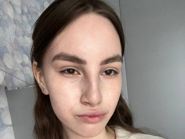 JinAvans has pale skin, brown eyes, dark eyebrows, and brown hair, with a slight pout and small, round nose. She's wearing a white shirt and has small earrings.