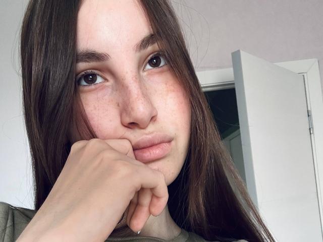 JinAvans has long brown hair, freckles on her face, and is resting her chin on her hand with a neutral expression. She has brown eyes and is wearing a green shirt.