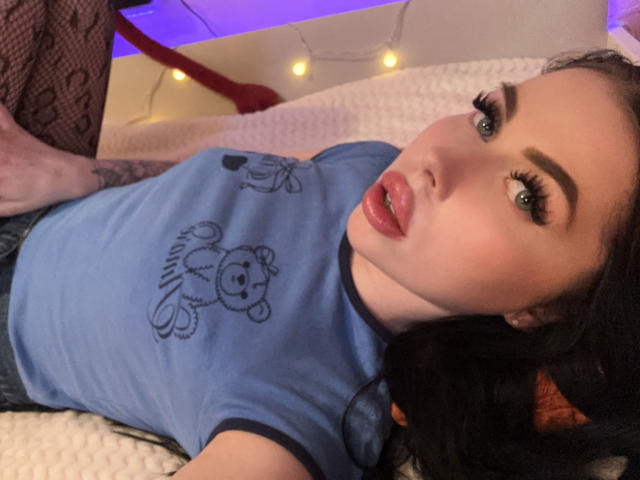 VelvetD with pouty lips, blue shirt, teddy bear print, fishnet legs, black hair, eyeliner, and tattooed arm, lying on white bed, looking at camera. Purple light in background.