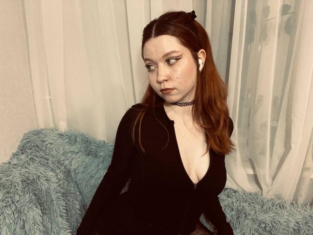 LilliamChevalier has pale skin, red hair, black choker, and deep cleavage in a black dress, sitting on a blue fuzzy couch with white curtains in the background.
