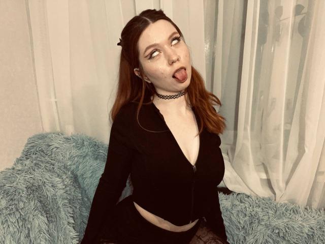 LilliamChevalier has pale skin, red hair, and a black choker, sticking her tongue out, wearing a black long-sleeve top, and sitting on a blue furry couch.