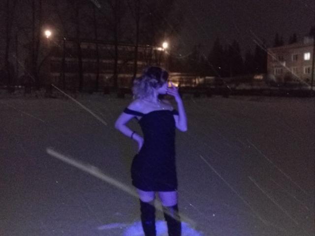 IngerKennington stands in a dark field at night, wearing a black off-shoulder dress and thigh-high socks, holding a phone to her ear, with her ass slightly raised.