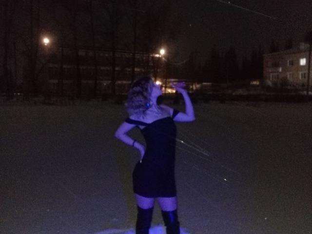 IngerKennington stands in dark, wearing a black off-shoulder dress and thigh-high socks, pissing into the air with one hand on her hip, in a nighttime photo.