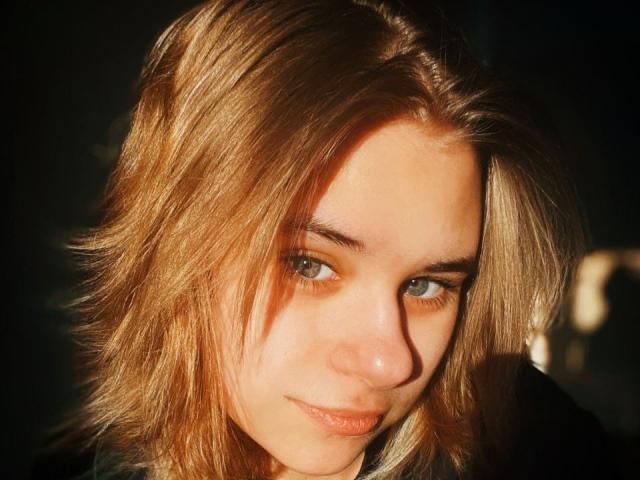 IngerKennington has dirty blonde, wavy hair, light skin, and green eyes with a slight smirk. Her face is illuminated by warm sunlight, highlighting her soft features.