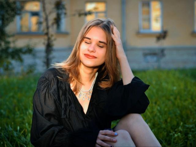 IngerKennington with blonde hair, eyes closed, wearing a black dress and pearl necklace, sitting on grass, hand in hair, smiling softly, showing her tits slightly.