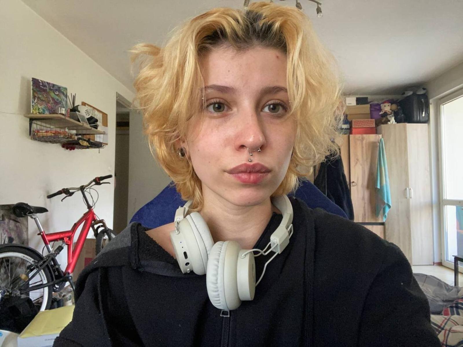 ShalonFregia with messy blonde hair, green eyes, nose ring, and white headphones, pouting her lips, wearing a black hoodie, in a cluttered room with a red bike.