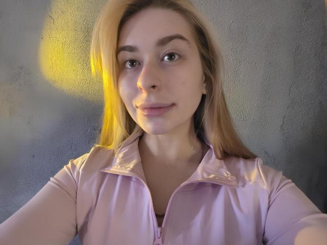 Maryln Mancinelli, con cabello rubio, una camisa rosa claro con cremallera y una sonrisa sutil, mostrando su rostro y parte superior del pecho, iluminada por una luz amarilla. Sus senos son pequeños y no son visibles..
