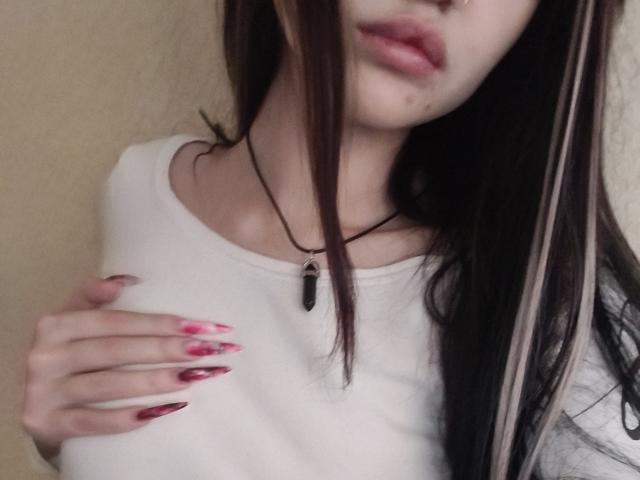 MistyEllstrom with long brown hair, pink-nailed hand on white shirt, black necklace with black gem, soft pink lips, slight pimple on chin, blurred upper face.