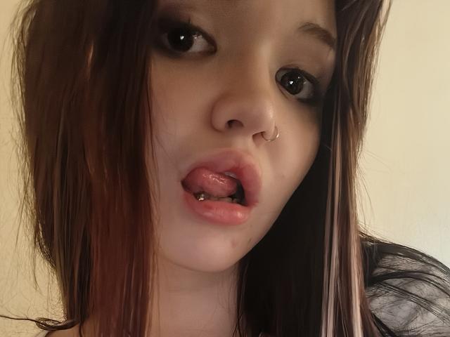 MistyEllstrom's face with brown hair, nose ring, and tongue out, showing a metal tongue piercing. She has dark eyes and pink, slightly parted lips.