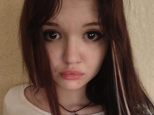 MistyEllstrom has brown eyes, a nose ring, and pink lips. She's wearing a white shirt with long brown hair and a black necklace. Her face is slightly pouty.