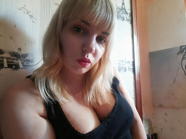 Leylisse has blonde hair, red lipstick, and is wearing a black tank top that shows her big, shiny tits. She's looking at the camera with a serious expression.