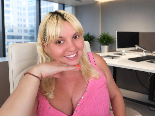 Leylisse with blonde hair, pink sleeveless top showing her tits, smiling at the camera, hand on chin, orange nail polish, bracelet, in an office with a white chair.