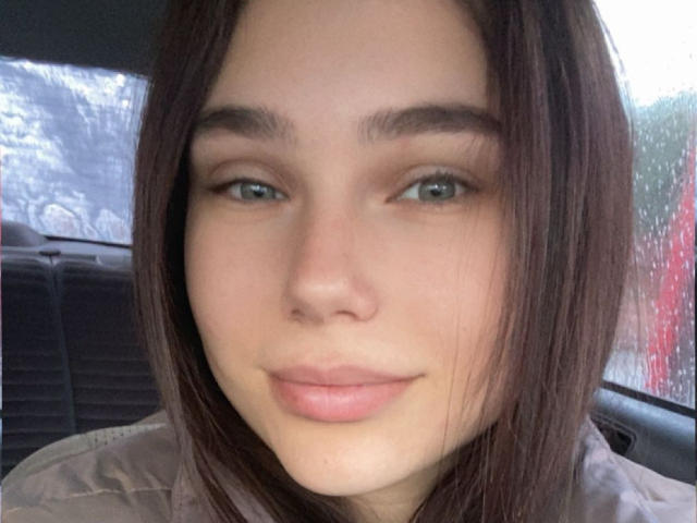 AnnaBellas hat glattes, braunes Haar, grüne Augen und ein leichtes Lächeln. Ihre rosafarbenen Lippen sind entspannt. Sie trägt eine hellgraue, gesteppte Jacke in einem Auto..