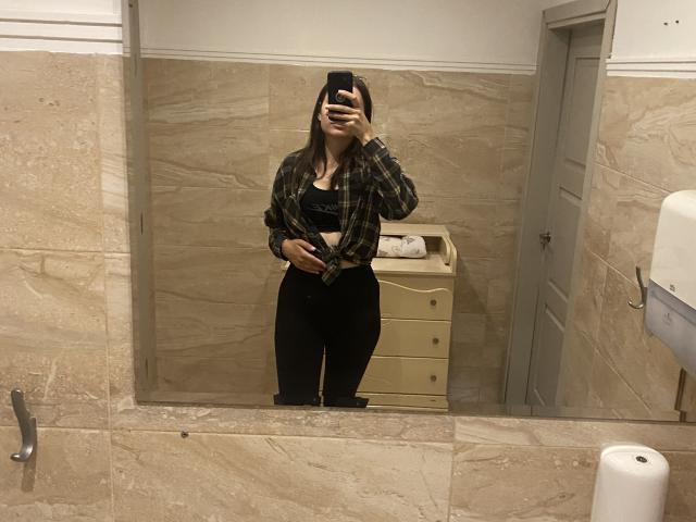 AnnaBella mit langen, braunen Haaren, die ein schwarzes Oberteil, ein kariertes Hemd und eine schwarze Hose trägt, steht in einem Badezimmer und macht ein Selfie vor dem Spiegel mit ihrem Handy, wobei sie ihre Genitalien bedeckt..
