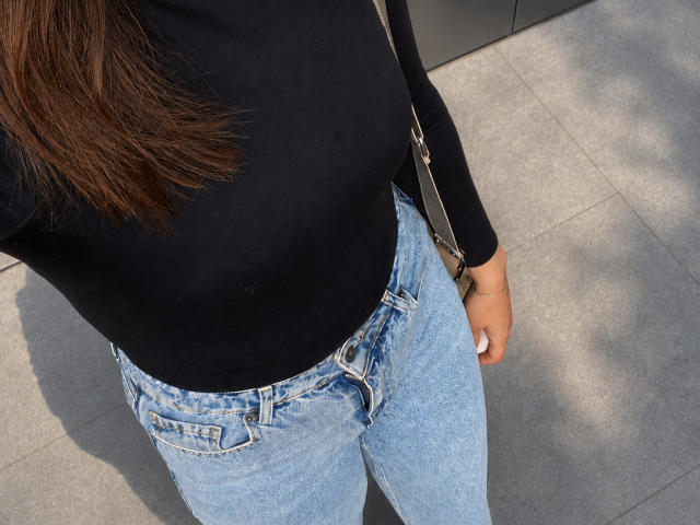 AnnaBellas has brown hair, a black top, and blue jeans. Her ass is covered by the jeans. She's holding a silver bag strap. Her hand is visible.