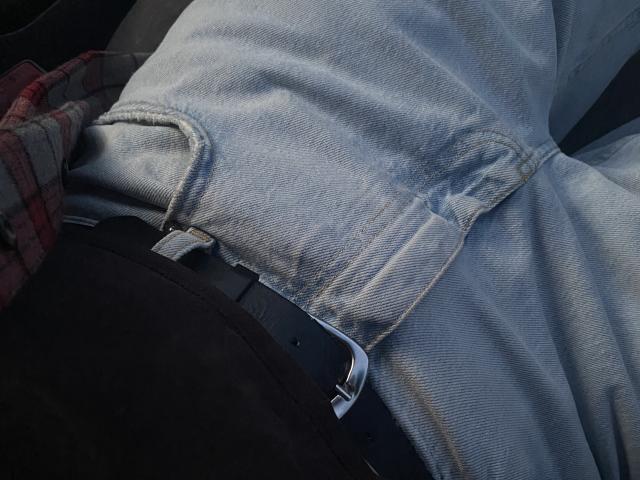 I jeans blu leggermente usurati di AnnaBellas, con una cintura nera, seduta, che mostra la texture del denim e la fibbia metallica della cintura, primo piano. La sua camicia a quadri rossa è parzialmente visibile..