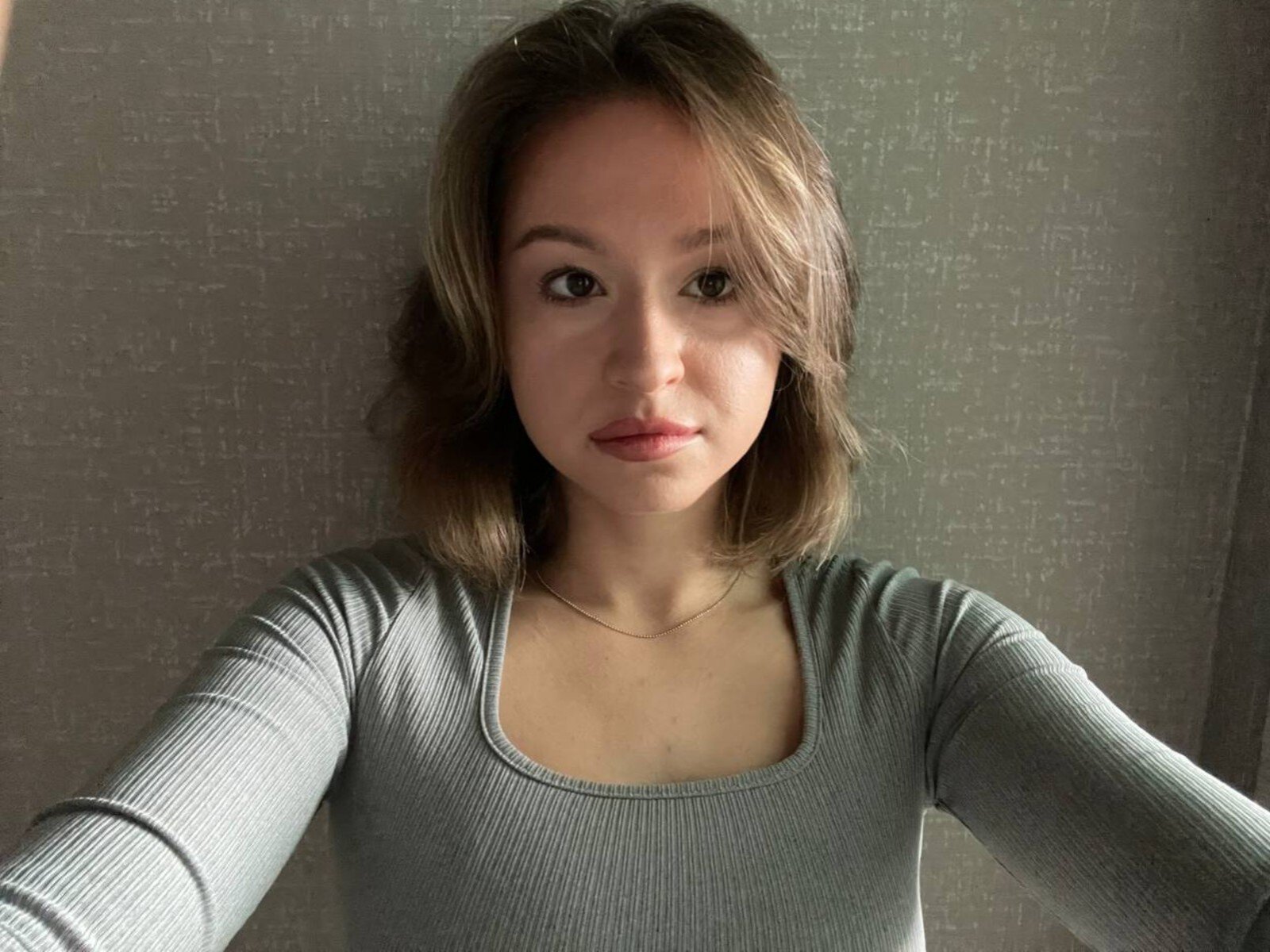 QuyenSaenger, con un seno di media grandezza, indossa una maglia a maniche lunghe grigia a coste, ha capelli castano chiaro ondulati e un sorriso discreto, ed è in piedi di fronte a una parete grigia con una texture particolare..
