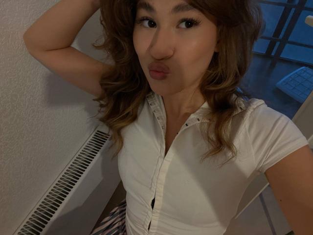 SerenaHuseth, pouting with her lips slightly pursed, wearing a white button-up shirt, her wavy brown hair falling to her shoulders, standing in a dimly lit room.