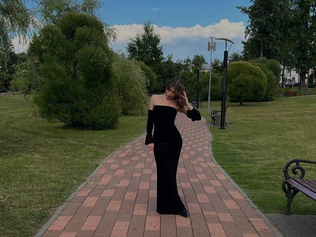 SerenaHuseth stands on a brick path, wearing a black, off-shoulder dress, her ass slightly accentuated, looking down with wavy brown hair, in a park.