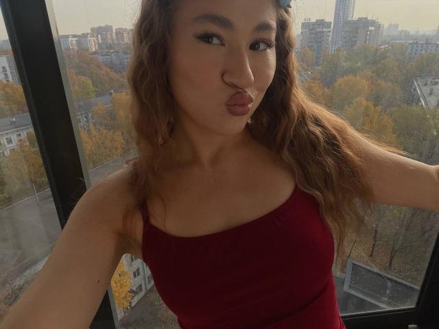 SerenaHuseth with pouty lips, wearing a red tank top, wavy brown hair, and black eyeliner, standing by a window with cityscape and autumn trees outside.