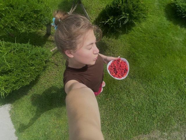 SerenaHuseth with brown top, blonde hair in buns, eating from white bowl of red berries, holding out arm for selfie, grassy yard, small bushes in background.