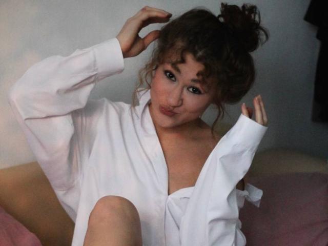 SerenaHuseth with brown curly hair in a bun, wearing a white shirt, pouting with slightly parted lips, one hand in her hair, and the other near her chest.