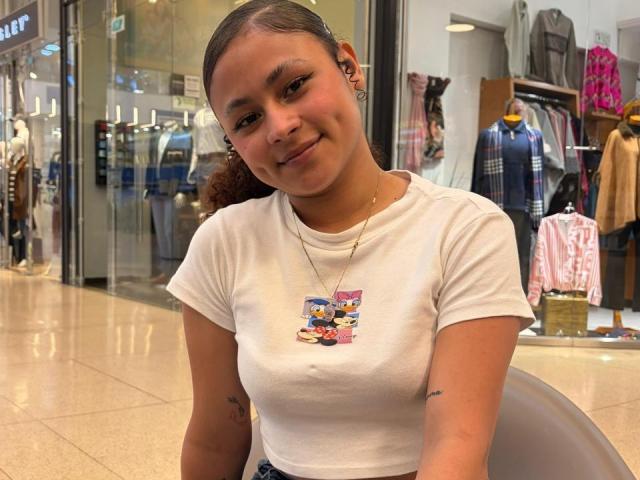 SofiaPassionne with light brown skin, curly dark hair, small tits in a white tee with a cartoon, smiling, wearing a gold necklace, sitting in a mall.