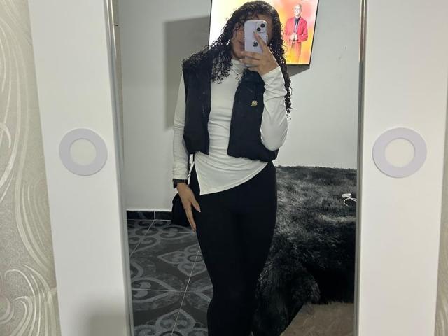 SofiaPassionne with curly hair, white long-sleeve shirt, black vest, and black pants, taking a mirror selfie, holding a phone, showing a slim, curvy ass.